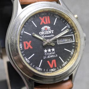以下が含まれることがあります： 黒い文字盤、赤いローマ数字、白い針を備えた、シルバーカラーのオリエント自動巻き腕時計。 茶色の革ストラップと日付表示が付いています。 文字盤には「Crystal 21 Jewels」と書かれています。