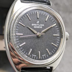 May include: A vintage Breitling Genève watch with a black dial and silver hands and markers. The watch has a stainless steel case and a black leather strap. The dial features the Breitling logo and the words "Breitling Genève". The watch is marked "Swiss Made".