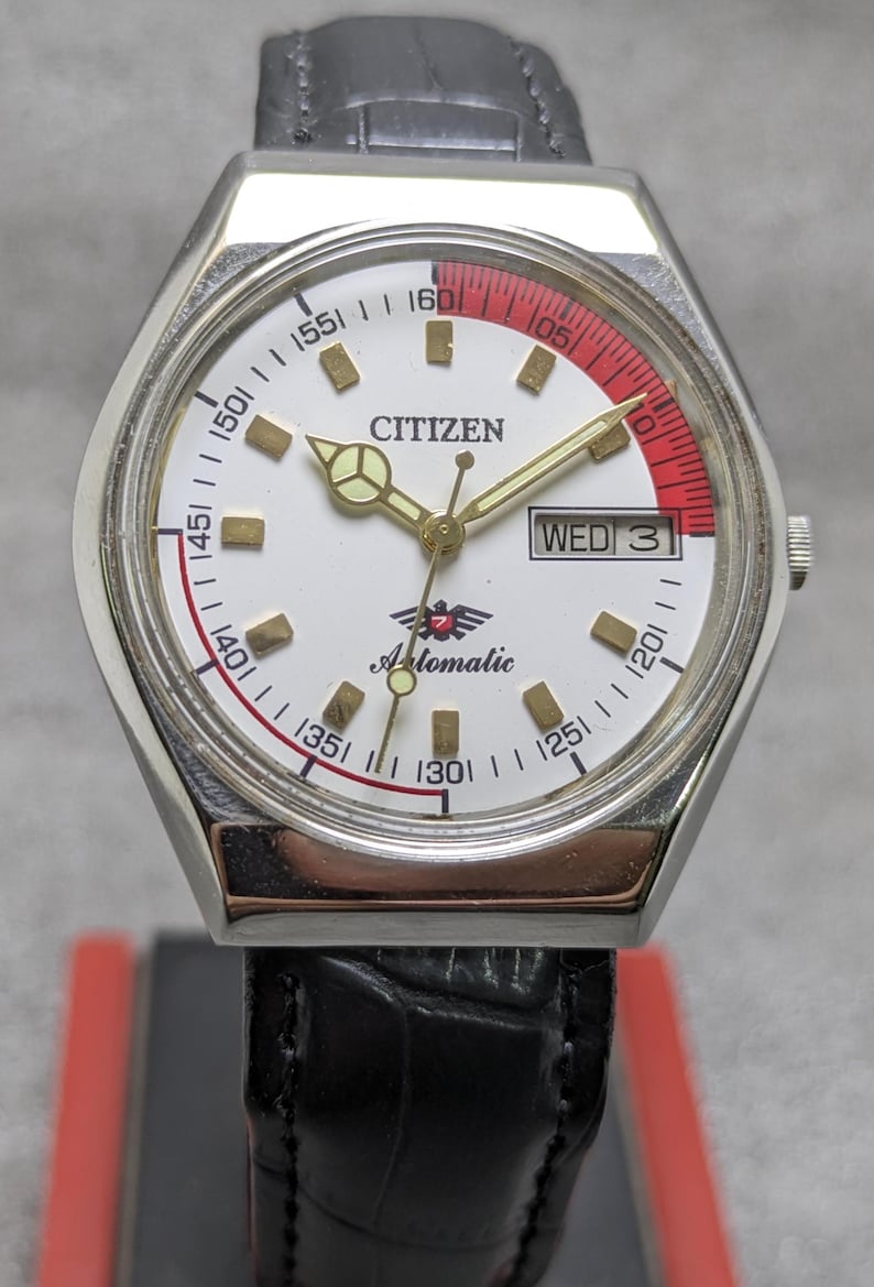 Puede incluir: Un reloj de pulsera autom&aacute;tico Citizen vintage con esfera blanca, agujas doradas y un acento rojo y blanco. La correa de cuero negro complementa la esfera, que muestra el d&iacute;a. La esfera incluye las palabras "Citizen" y "Automatic".