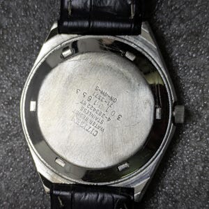 Puede incluir: Primer plano de la parte trasera de un reloj Citizen de acero inoxidable con una correa de cuero negro. La esfera del reloj es plateada con texto grabado, incluyendo "Citizen Water Resist Stainless". El reloj est&aacute; sujeto a una correa de cuero negro.