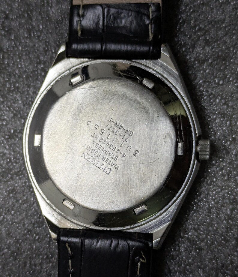 Puede incluir: Primer plano de la parte trasera de un reloj Citizen de acero inoxidable con una correa de cuero negro. La esfera del reloj es plateada con texto grabado, incluyendo "Citizen Water Resist Stainless". El reloj est&aacute; sujeto a una correa de cuero negro.