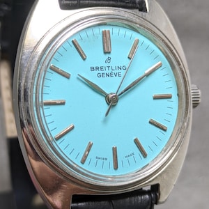 May include: A vintage Breitling Genève watch featuring a light blue face with gold-tone hands and markers. The watch has a silver-tone case and a black leather strap. The words "Breitling Genève" and "Swiss Made" are visible.