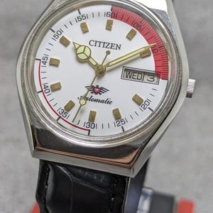 Puede incluir: Un reloj de pulsera autom&aacute;tico Citizen con esfera blanca, agujas doradas y un acento rojo. El reloj tiene una correa de cuero negro y muestra el d&iacute;a de la semana. La esfera incluye la palabra "Citizen" y la palabra "Automatic".