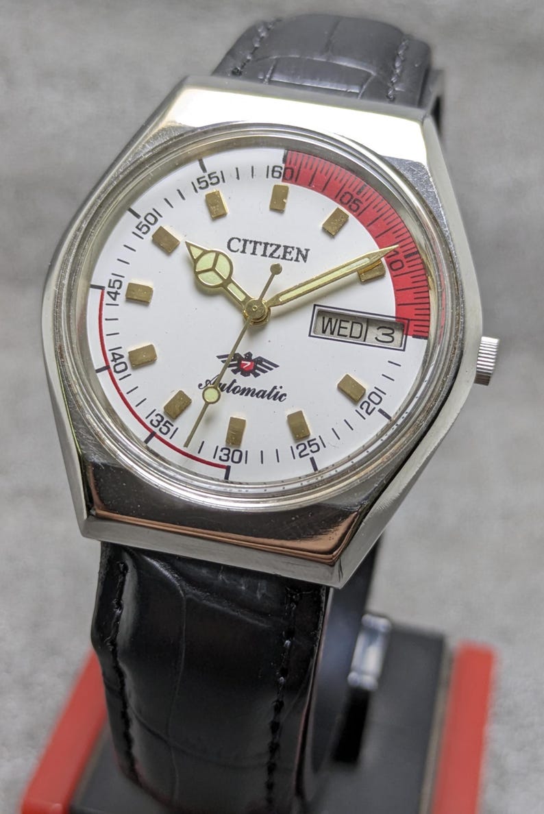 Puede incluir: Un reloj de pulsera autom&aacute;tico Citizen con esfera blanca, agujas doradas y un acento rojo. El reloj tiene una correa de cuero negro y muestra el d&iacute;a de la semana. La esfera incluye la palabra "Citizen" y la palabra "Automatic".