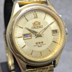 May include: Gold-toned Orient watch with a gold dial, featuring the day and date. The watch face displays the word "Orient" and "Water Resist 50m". The watch has a gold-colored band and a gold bezel.