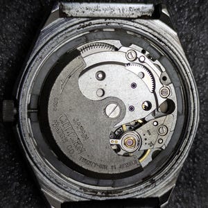 Puede incluir: Primer plano de la parte trasera de un reloj Citizen, que revela el intrincado movimiento mec&aacute;nico. El mecanismo del reloj presenta engranajes plateados, un volante y el texto "Citizen Watch Co. Japan Twenty-One 21 Jewels".
