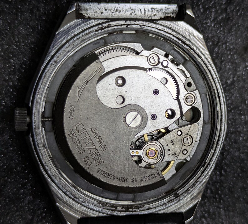 Puede incluir: Primer plano de la parte trasera de un reloj Citizen, que revela el intrincado movimiento mec&aacute;nico. El mecanismo del reloj presenta engranajes plateados, un volante y el texto "Citizen Watch Co. Japan Twenty-One 21 Jewels".
