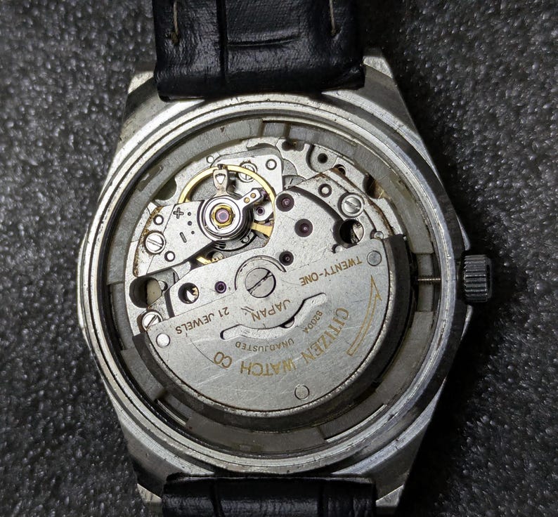 Może przedstawiać: Zbliżenie zegarka Citizen bez tylnej części, ukazujące skomplikowany mechanizm. Mechanizm zegarka zawiera liczne małe koła zębate, kamienie i wirnik. Zegarek ma czarny sk&oacute;rzany pasek.