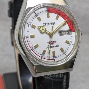 Puede incluir: Un reloj de pulsera autom&aacute;tico Citizen vintage con esfera blanca, agujas doradas y un acento rojo. El reloj cuenta con una pantalla de d&iacute;a y fecha y est&aacute; montado en una correa de cuero negro. La esfera del reloj muestra la palabra "Citizen" y "Automatic".