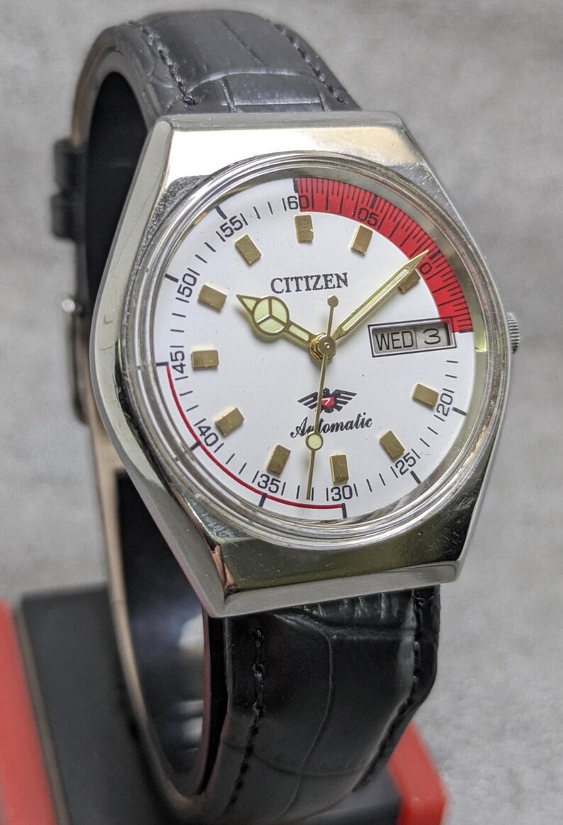 Puede incluir: Un reloj de pulsera autom&aacute;tico Citizen vintage con esfera blanca, agujas doradas y un acento rojo. El reloj cuenta con una pantalla de d&iacute;a y fecha y est&aacute; montado en una correa de cuero negro. La esfera del reloj muestra la palabra "Citizen" y "Automatic".