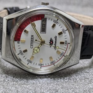 Puede incluir: Un reloj de pulsera autom&aacute;tico Citizen plateado con esfera blanca, manecillas doradas y correa de cuero negro. La esfera del reloj presenta un acento rojo y blanco, visualizaci&oacute;n del d&iacute;a y la palabra "Citizen".