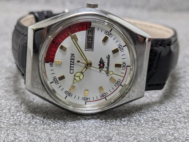 Puede incluir: Un reloj de pulsera autom&aacute;tico Citizen plateado con esfera blanca, manecillas doradas y correa de cuero negro. La esfera del reloj presenta un acento rojo y blanco, visualizaci&oacute;n del d&iacute;a y la palabra "Citizen".
