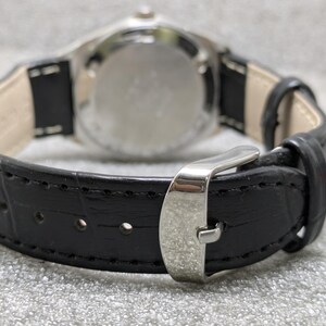Puede incluir: Primer plano de un reloj de pulsera con una correa de cuero negro y una hebilla plateada. La esfera del reloj es visible en el fondo. La correa tiene un patr&oacute;n texturizado y est&aacute; cosida a lo largo de los bordes.