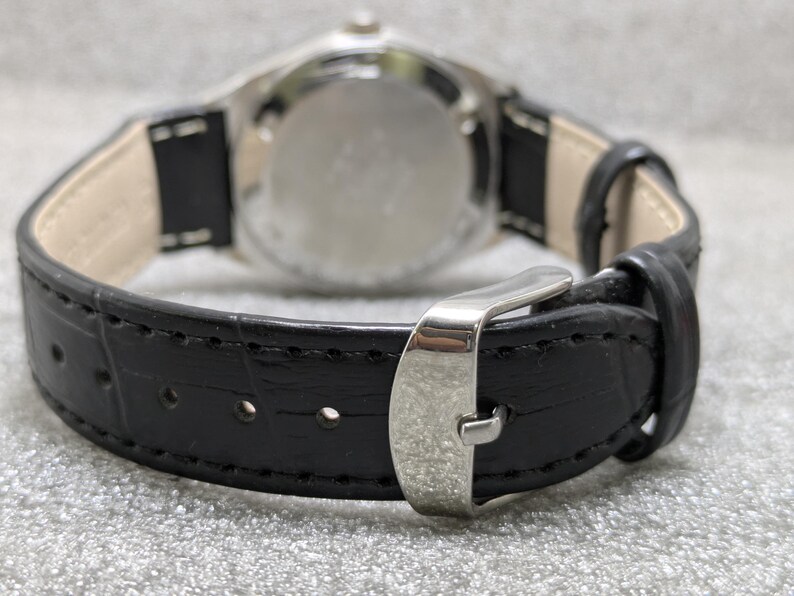 Puede incluir: Primer plano de un reloj de pulsera con una correa de cuero negro y una hebilla plateada. La esfera del reloj es visible en el fondo. La correa tiene un patr&oacute;n texturizado y est&aacute; cosida a lo largo de los bordes.