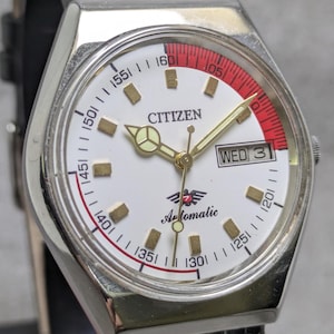 Puede incluir: Un reloj de pulsera autom&aacute;tico Citizen con esfera blanca, manecillas doradas e indicadores de hora dorados. El reloj tiene un acento rojo y una pantalla de d&iacute;a y fecha. El reloj est&aacute; en una correa de cuero negro.