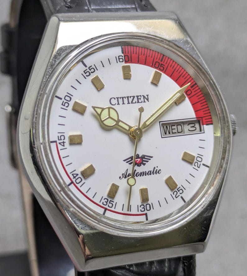 Puede incluir: Un reloj de pulsera autom&aacute;tico Citizen con esfera blanca, manecillas doradas e indicadores de hora dorados. El reloj tiene un acento rojo y una pantalla de d&iacute;a y fecha. El reloj est&aacute; en una correa de cuero negro.