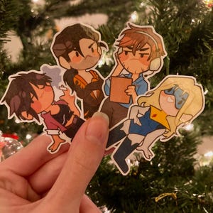 Puede incluir: Una mano sosteniendo cuatro pegatinas de personajes de dibujos animados. Las pegatinas presentan varios personajes con diferentes atuendos y expresiones. El fondo es un árbol de Navidad borroso con luces.