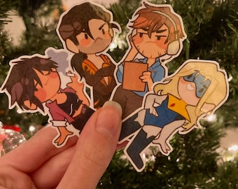 Könnte beinhalten: Eine Hand hält vier Aufkleber mit Cartoon-Charakteren. Die Aufkleber zeigen verschiedene Charaktere mit unterschiedlichen Outfits und Ausdrücken. Der Hintergrund ist ein verschwommener Weihnachtsbaum mit Lichtern.