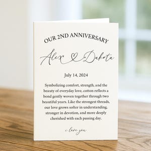 May include: A cream-colored anniversary card with the text "OUR 2ND ANNIVERSARY" and the names "Alex & Dakota." The date "July 14, 2024" is also printed. The card is on a wooden surface.