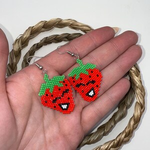 May include: A pair of beaded strawberry earrings. The earrings feature a red body with black seed details, green tops, and white smiles. The earrings are hanging from silver hooks.