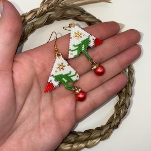 May include: Handmade Christmas tree earrings. The earrings feature a white beaded tree design with green accents, red beaded tips, and a gold snowflake. A red bead hangs from the bottom of each earring. The earrings are held by a hand.