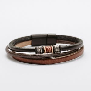 Puede incluir: Un brazalete de cuero multicapa con cierre magnético. El brazalete presenta una correa de cuero marrón, negro y gris con una cuenta decorativa en el centro.