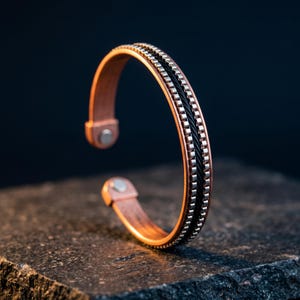 May include: A copper-toned cuff bracelet featuring a central black twisted cable inlay flanked by two rows of small silver-toned square metallic studs. The open-ended design includes a circular metallic element at each terminal end of the band.