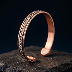 Pode incluir: Uma pulseira tipo bracelete em tom de cobre com uma incrustação central de corrente em tom prateado. A pulseira tem um acabamento liso e polido nas bordas externas e inclui um pequeno íman circular embutido na extremidade interna.