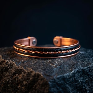 May include: A cuff bracelet featuring a central twisted copper wire flanked by two smooth black bands, all encased within a polished copper frame. The open-ended design includes rounded copper terminals at each end of the metallic structure.