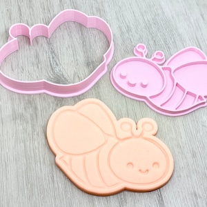May include: Two pink plastic cookie cutters, one shaped like a bee and the other shaped like a cloud. The bee cutter has a simple design with two small circles for eyes and a line for a mouth. The cloud cutter has a rounded shape with a slightly textured surface.
