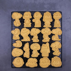 May include: A set of 20 yellow cookie cutters featuring various characters from the Peanuts comic strip, including Snoopy, Charlie Brown, Lucy, and Woodstock. The cutters are arranged on a black wire rack.