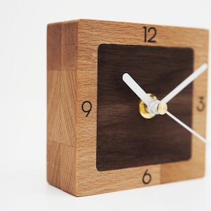 Op de afbeelding: Een vierkante houten klok met een lichtbruine lijst en een donkerbruin midden. De wijzerplaat heeft witte wijzers en zwarte cijfers op de posities 3, 6, 9 en 12 uur. De klok is een decoratief item.