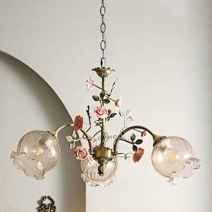 May include: A vintage-style chandelier with three glass globe shades and delicate pink rose accents. The fixture has a bronze finish and is suspended from a chain. A matching wall sconce is also visible, adding to the romantic ambiance.