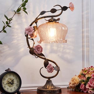 Puede incluir: Una lámpara de mesa decorativa con un diseño floral. La lámpara presenta una pantalla de cristal rosa y está adornada con rosas rosas y enredaderas de metal color bronce. También se ve un reloj analógico negro y un ramo de flores.