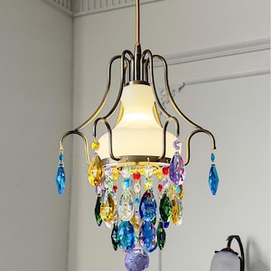 Pode incluir: Um candelabro de estilo vintage com uma estrutura de cor bronze e uma sombra de vidro fosco. O candelabro é adornado com pingentes de cristal coloridos e facetados em tons de azul, amarelo, verde e roxo, criando uma luminária vibrante e elegante.