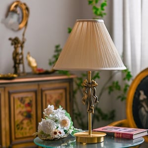 May include: A table lamp with a brass-toned metal base featuring a decorative bow detail on the stem and a pleated, light beige fabric lampshade. The lamp has a circular base and a slender vertical post supporting the shade.