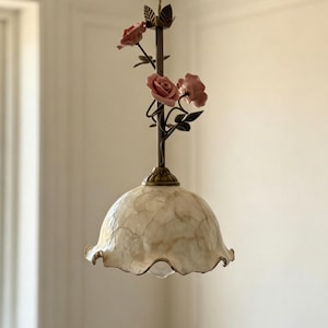 May include: A decorative pendant light with a floral design. The light fixture features a cream-coloured, shell-like shade with a scalloped edge. Pink rose accents and bronze-coloured metal details add to the fixture's elegant aesthetic. The light hangs from a twisted cord.