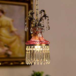 May include: A hanging pendant light fixture featuring a deep pink fluted glass shade, an ornate antique brass metal frame with scrollwork, and multiple clear teardrop-shaped crystal prisms dangling from the top and bottom edges.