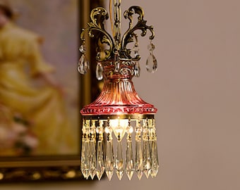 May include: A hanging pendant light fixture featuring a deep pink fluted glass shade, an ornate antique brass metal frame with scrollwork, and multiple clear teardrop-shaped crystal prisms dangling from the top and bottom edges.
