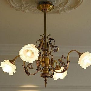 May include: A gold chandelier with five white glass flower-shaped shades. The chandelier has a decorative, ornate design with intricate details.