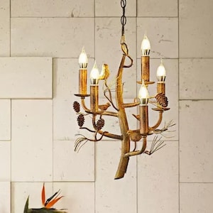 May include: A gold-colored, tree-branch-shaped chandelier with six candle-style lights. The chandelier features small bird and pinecone accents. The fixture hangs above a red cabinet with a vase of flowers and a framed picture.