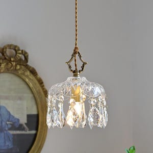 May include: A clear glass chandelier pendant light with crystal drops hanging from the bottom. The light is suspended from a brown cord and has a brass finish.