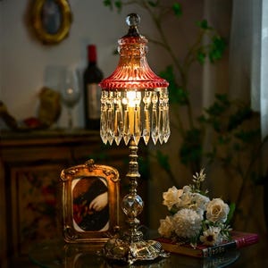 Puede incluir: Una lámpara de mesa de estilo vintage con una pantalla de vidrio roja y detalles de cristal. La lámpara tiene una base dorada y está iluminada por una bombilla de luz cálida.