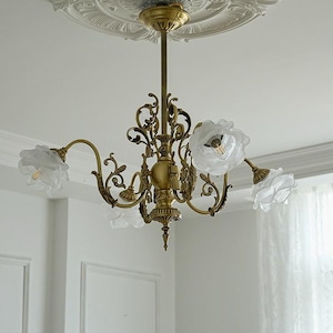 May include: A gold-toned chandelier with five white flower-shaped lampshades. The chandelier has a decorative, ornate design with intricate details.