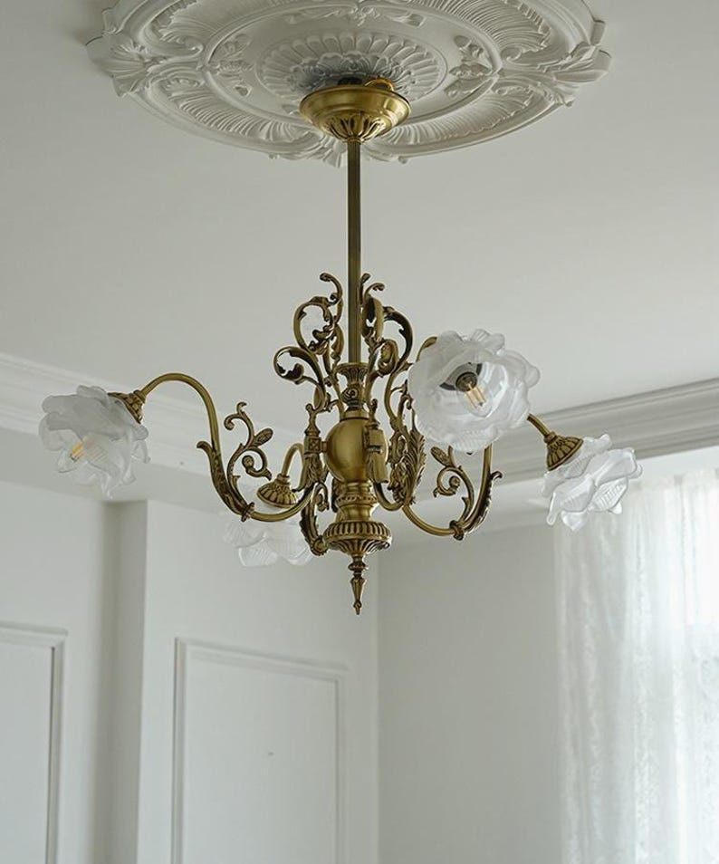 May include: A gold-toned chandelier with five white flower-shaped lampshades. The chandelier has a decorative, ornate design with intricate details.