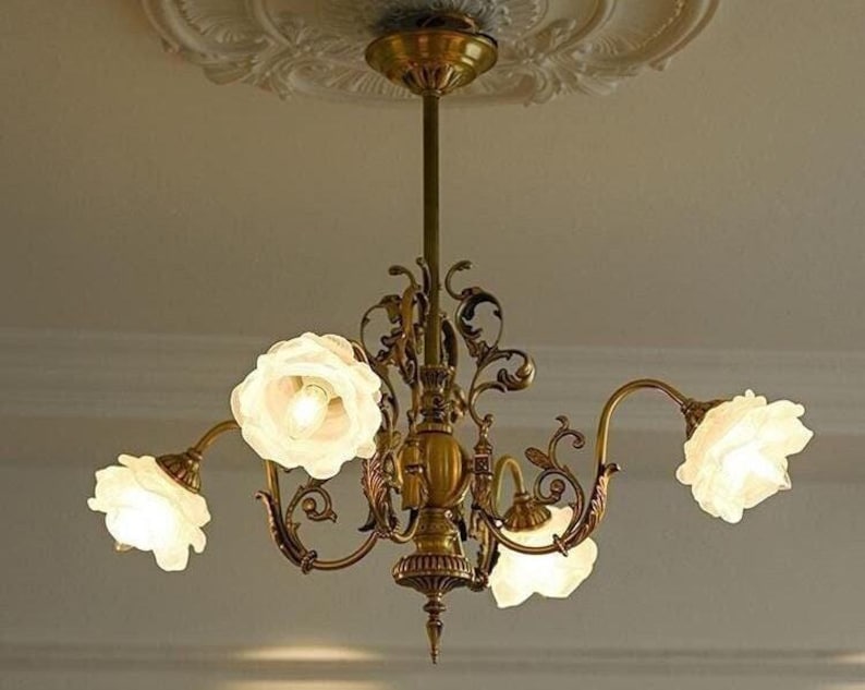 May include: A gold chandelier with five white glass flower-shaped shades. The chandelier has a decorative, ornate design with intricate details.