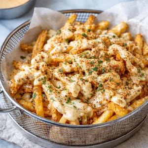 May include: A basket of golden-brown french fries, generously topped with creamy white cheese sauce and sprinkled with herbs and spices. The fries are served in a wire mesh basket lined with parchment paper.