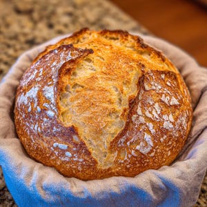 May include: A freshly baked loaf of sourdough bread, golden brown with a crispy crust, sits in a linen-lined basket. The bread's surface is dusted with flour, and the texture shows a rustic, artisan quality. The bread is round and has a beautiful, natural appearance.