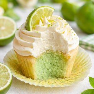 Op de afbeelding: Een limoen cupcake met witte frosting en een schijfje limoen. De cupcake heeft een groene binnenkant en er is een hap uit genomen. De cupcake zit in een gele papieren vorm, met limoenen op de achtergrond.