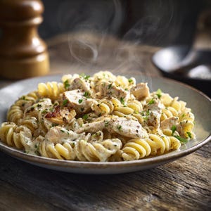May include: A close-up shot of a bowl of creamy pasta with chicken and herbs. The pasta is fusilli, and the chicken is cut into bite-sized pieces. Steam rises from the dish, suggesting it is freshly prepared. The bowl sits on a wooden surface.
